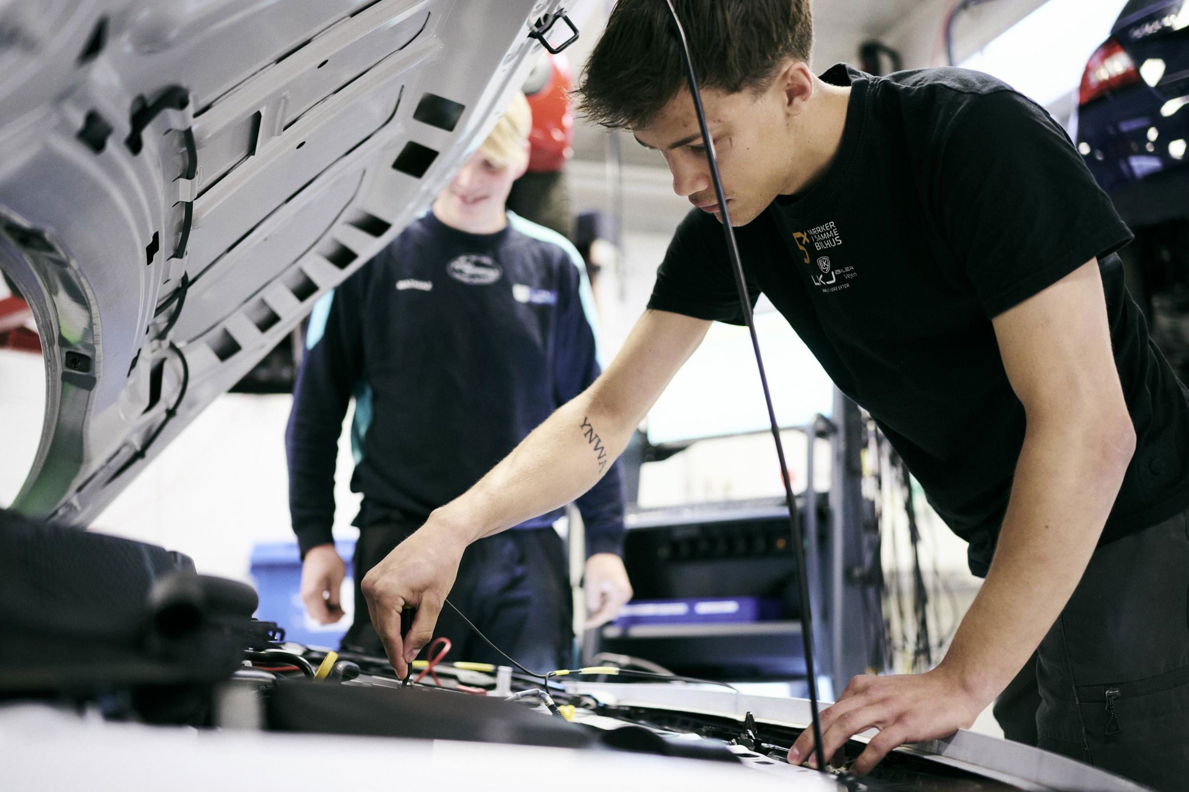 To personvognsmekanikerlærlinge fejlsøger på en bilmotor på skolens autoværksted.
