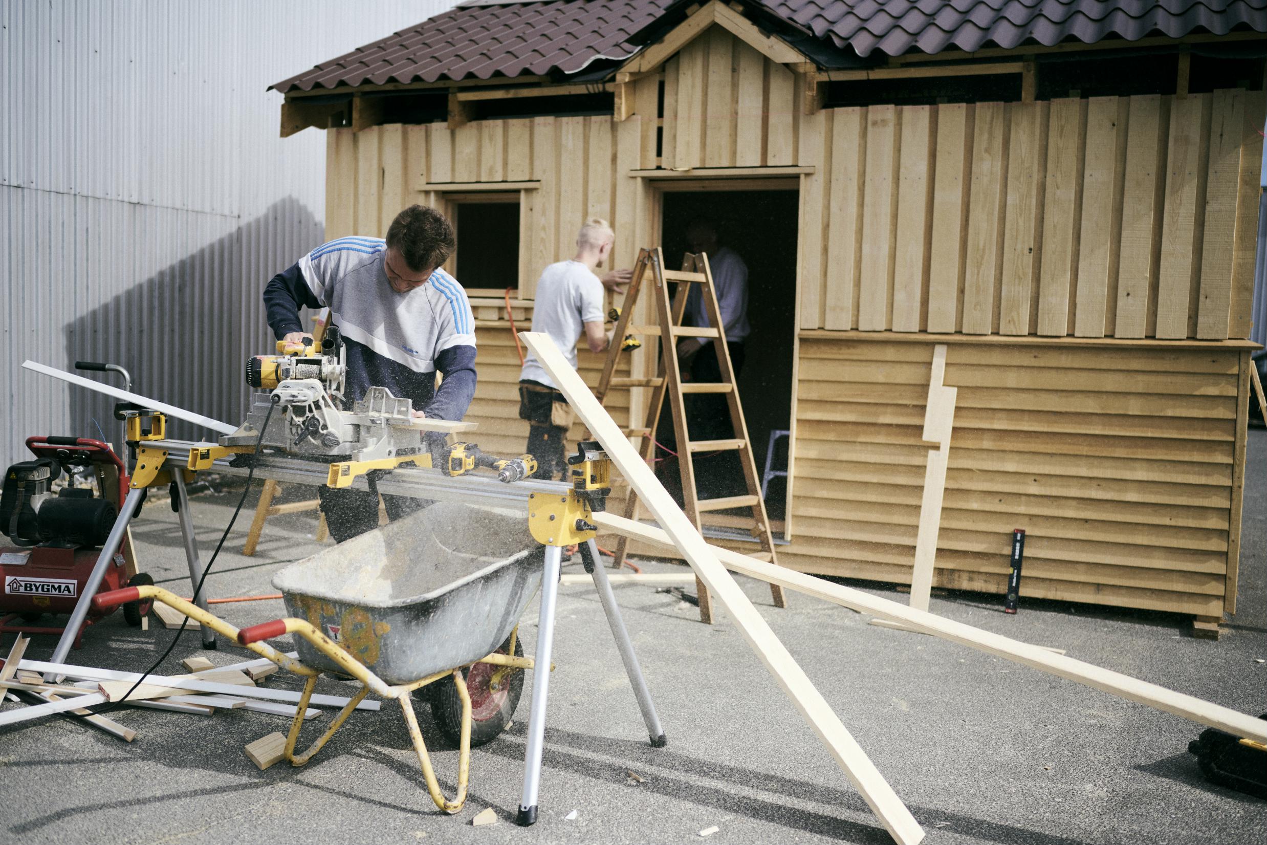 En tømrerelev bruger rundsaven, mens to andre elever bygger på et annekshus i Hansenbergs skoleoplæring.