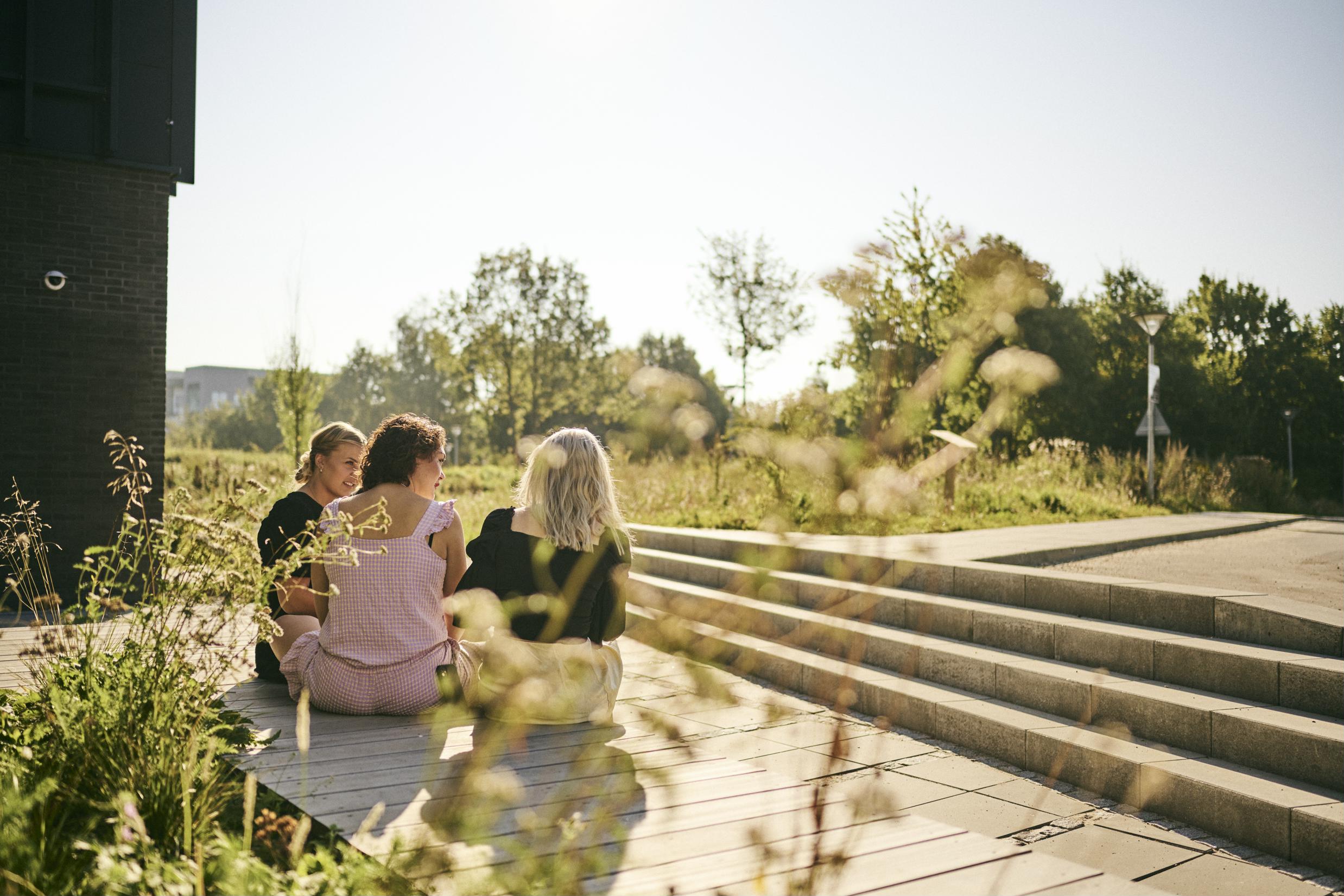 3 frisørelever sidder en sommerdag udenfor Hansenberg og hygger med udsigt til et grønt område med biodiversitet.