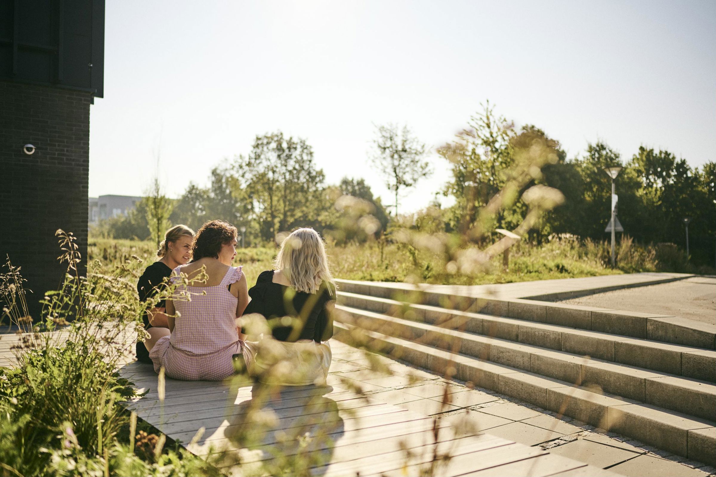 3 frisørelever sidder en sommerdag udenfor Hansenberg og hygger med udsigt til et grønt område med biodiversitet.